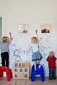 Three children scribbling on a white wall with framed photographs, plastic chairs and dolls' house