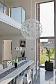 Spherical pendant lamp with white flower details above modern, white dining area with glossy floor below gallery with glass balustrade