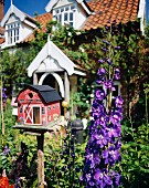 Bunt bemaltes Vogelhäuschen neben lila Rittersporn vor englischem Landhaus