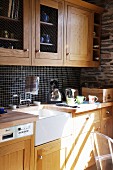 Einbauküche mit Holzfronten und Porzellanspüle; schwarze Mosaikfliesen und rustikale Natursteinwand im Hintergrund