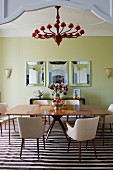 Large, square, exotic wood dining table, retro chairs with pale upholstery, red chandelier and framed mirrors on painted wall in spacious dining room