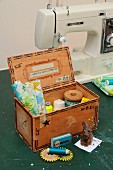 Upcycled cigar box decorated with stamped prints and washi tape repurposed as sewing box containing fabric remnants and reels of thread on work table in front of sewing machine