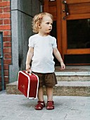 Blonde little girl holding red suitcase waiting outside front door