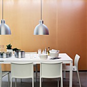 Crockery on white table below pendant lamps with metal lampshades in front of pale brown panel wall