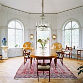 Long dining table, Biedermeier chairs and chandelier in white, wood-panelled dining room with arched windows
