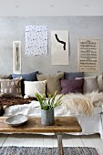 Shabby chic seating area with furs and scatter cushions on sofa; artistic pages pinned on concrete wall and jar of narcissus o vintage bench used as coffee table
