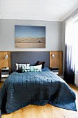 Double bed with dark blue spread and fabric and wooden headboard panel on wall below photo of desert in traditional bedroom