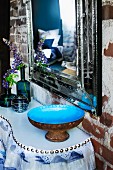 Blue-glazed bowl and smoked glass vases of lupins on console table below antique mirror on brick wall