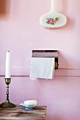 Candlestick on wooden table and floral bath brush above toilet roll holder on pink-painted wall