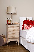 Shabby-chic, renovated bedside cabinet next to white bed with bright red cushions