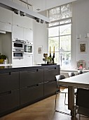 Modern kitchen with floor-to-ceiling window, white fitted cupboards and dark brown island counter in open-plan interior