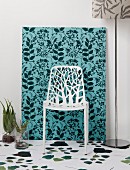 Delicate white chair in front of frame covered in jungle-patterned fabric and leaf pattern on white floor
