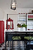 Barhocker aus schmiedeeisernem Gestell und grüne Sitzpolster vor Frühstücksbar, dahinter Wandspiegel mit rotem, verziertem Rahmen und weisser Vitrinenschrank