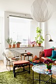 Corner of living room with stool, side tables, colourful tray, green flokati rug, spherical pendant lamp and classic, wire chair below window