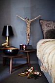 Bedside Gun table lamp and Jesus figurine on wooden bedside table with multiple round surfaces next to bed with fur blanket and sports shoes on floor