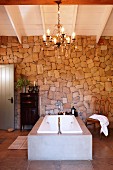 Free-standing bathtub with concrete surround below chandelier in front of stone wall in rustic setting