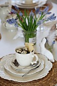 Easter place setting for afternoon tea with bird's nest and bunny biscuit in cup and white china bunny; grape hyacinths in jam jar in background
