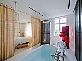 Free-standing bathtub in ensuite bathroom with view into bedroom through open curtains