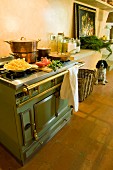 Copper pans on matt green, vintage cooker with brass rail in Tuscan kitchen-dining room
