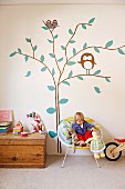 Child sitting in wicker shell chair with soft toys and rustic trunk against wall with tree mural