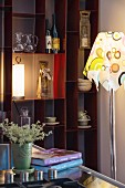 Standard lamp with retro lampshade in front of wine bottles and crockery on open-fronted shelves