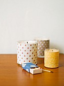 Tealight holders covered in patterned fabrics and box of matches on wooden surface