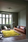 Minimalist living room painted sage green with brown leather sofa, bay window, designer rocking chair and green wool rug