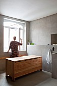 Designer Holzbadewanne mit geschlossenem Deckel auf Zementestrichboden in kühlem reduziertem Bad, Frau an geöffnetem Badfenster