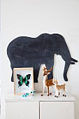 Butterfly in display case and small deer figurines on white cabinet below elephant-shaped blackboard
