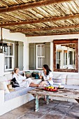 Two women chatting sitting on comfortable bench on roofed terrace with rustic coffee table on tiled floor