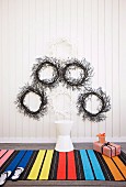 Wreaths in black and white on a white wooden wall, in front of it a colorful striped carpet with a stool and Christmas presents