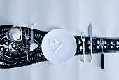 Love-heart of sugar sprinkles on white plate and cutlery with unusual handles next to pastry in heart-shaped dish on roll of wallpaper with black pattern