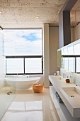Spacious designer bathroom with freestanding white bathtub below panoramic window, twin sinks and mirrored wall cabinet