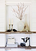 Vases, candlesticks and books on rustic wooden shelves built into niche with white-painted wooden back wall