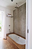 Concrete wall, rainfall shower and sunken white bathtub with stone-tiled surround in minimalist bathroom