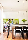 Fröhliches Kind mit Mutter vor weißer Designerküche und hellgrün spiegelndem Spritzschutz in offenem Wohnbereich