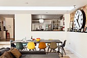 Blick vom Essbereich zur erhöten Küche mit Oberlicht über dem langen Tisch mit farblich unterschiedlichen Designerstühlen