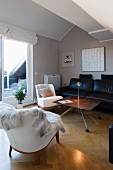 Fur blanket on white armchair and black leather sofa around DIY coffee table in attic interior with view of balcony