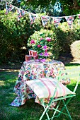 Festively decorated garden with bunting above table, floral tablecloth, cake stand of pastries and drinks; striped cushion on pastel green folding chair in foreground