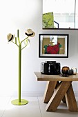 Various hats on green coat stand next to solid wooden table with various vases below framed picture on wall