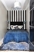 Quilted bedspread on double bed with black headboard against black and white striped wallpaper in narrow niche and rug on wooden floor at foot of bed