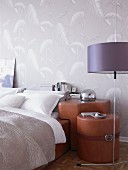 Detail of double bed with fitted, custom-made group of bedside cabinets covered in brown leather against wall with pale grey patterned wallpaper; standard lamp with purple lampshade in foreground
