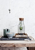 Nigella seed head in bottle with cork stopper in box next to bowl and ceramic storage jar on rustic wooden surface; vintage key hanging on marble wall in background