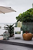 White wicker chair, large floor lantern and drum-shaped side table on terrace made from reclaimed wood and plastic with glass balustrade with sea view