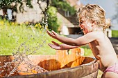 Kleines Mädchen in Badehose spritzt mit Wasser vor Holzfass
