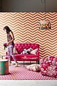 Seating area in feminine shades of pink with antique sofa, wallpaper and rug in zigzag patterns, floral patterned armchair and lampshade and woman wearing mixture of patterns