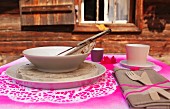 Place setting with soup bowl and wooden cutlery tied to linen napkin on place mat sprayed hot pink through doily stencils in front of alpine cabin