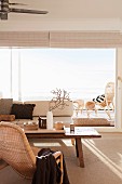 Wicker armchair, pale sofa, rustic coffee table and panoramic window in bright, pleasant living room