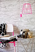 Toiletries on chrome side table next to stacked towels on chair against stone-effect wallpaper and below pendant lamp with pink wire lampshade