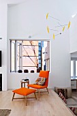 Orange chair and matching footstool on platform in front of window-like opening in wall; mobile hanging from ceiling